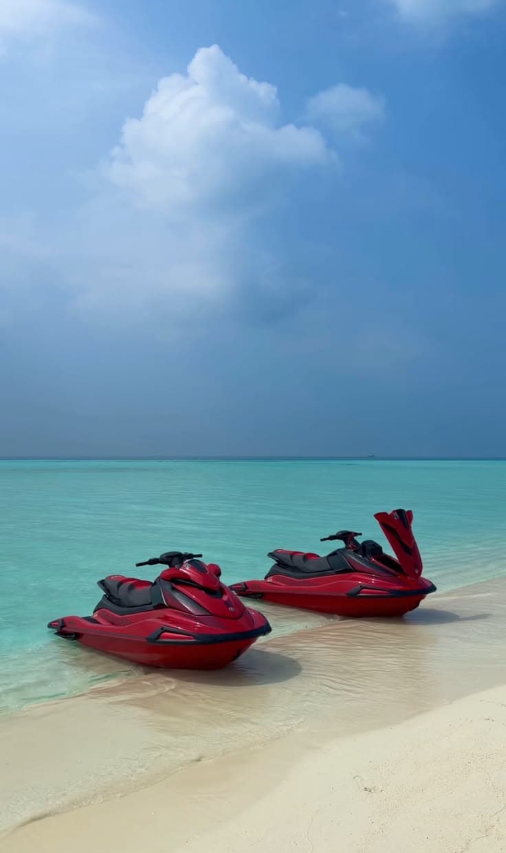 Two red jet skis parked on a white sand beach beside clear turquoise tropical water.