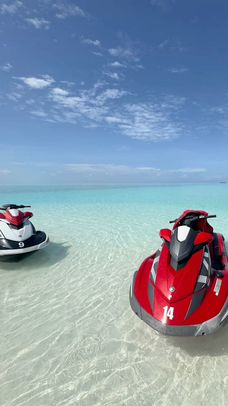 Two jet skis floating in shallow, crystal clear turquoise water under a bright blue sky.